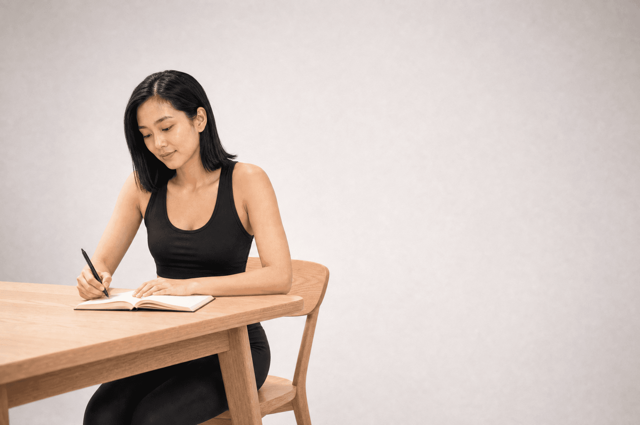 A woman in black loungewear writing in a notebook at a pale wood desk in a white studio