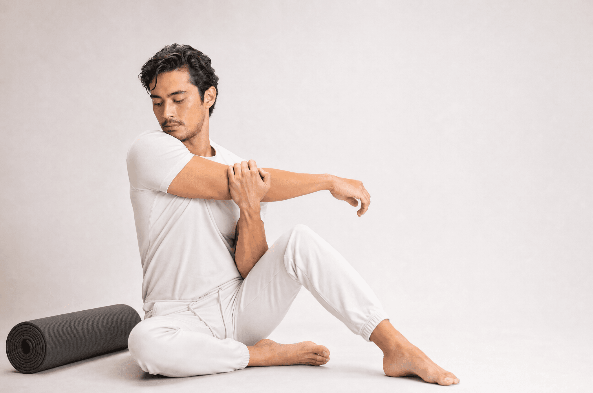 A man in white athleisure seated on a studio floor stretching one shoulder, a rolled grey mat beside him