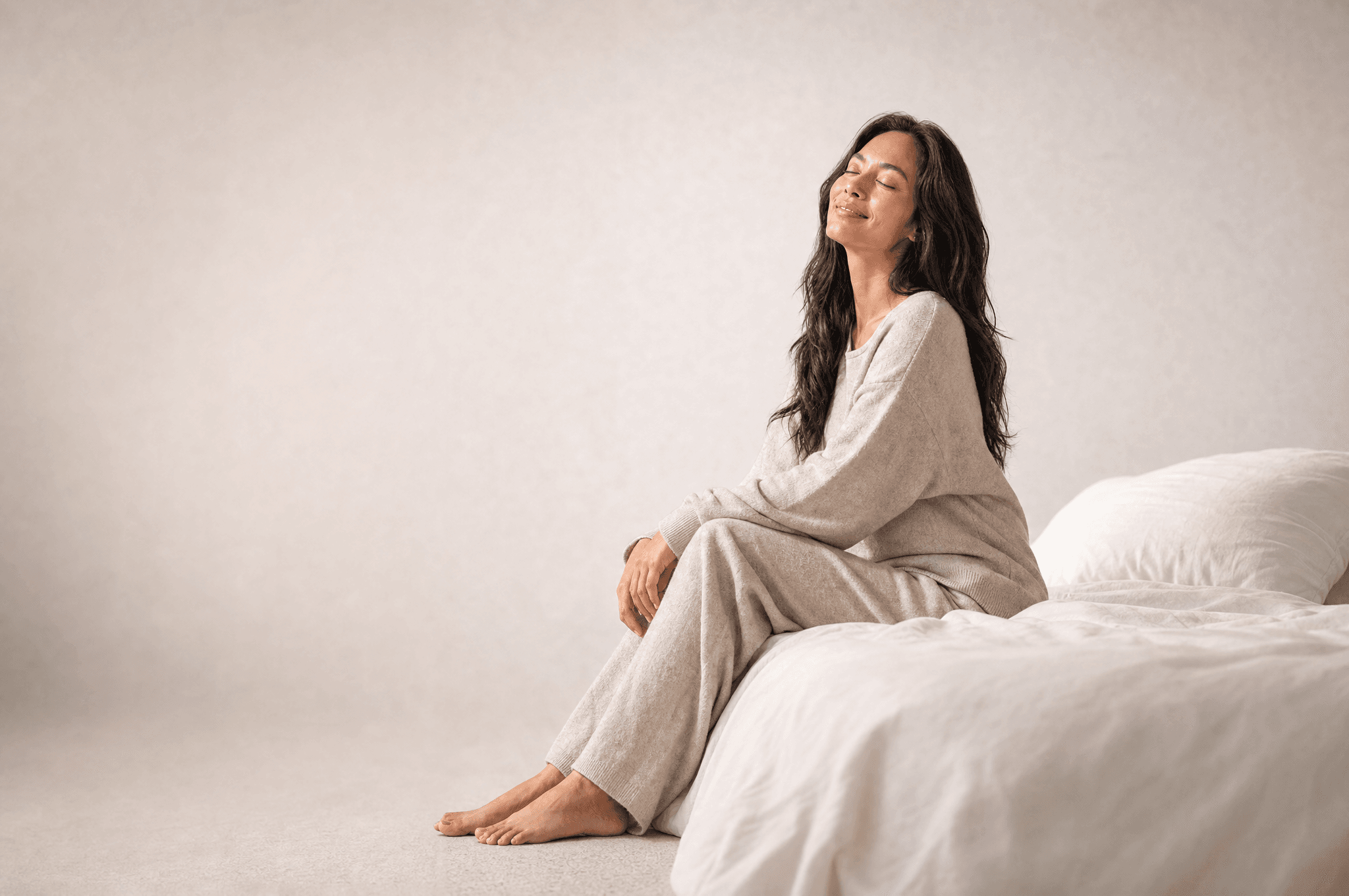 A woman in oat loungewear sitting on the edge of a white bed, eyes closed, face turned up