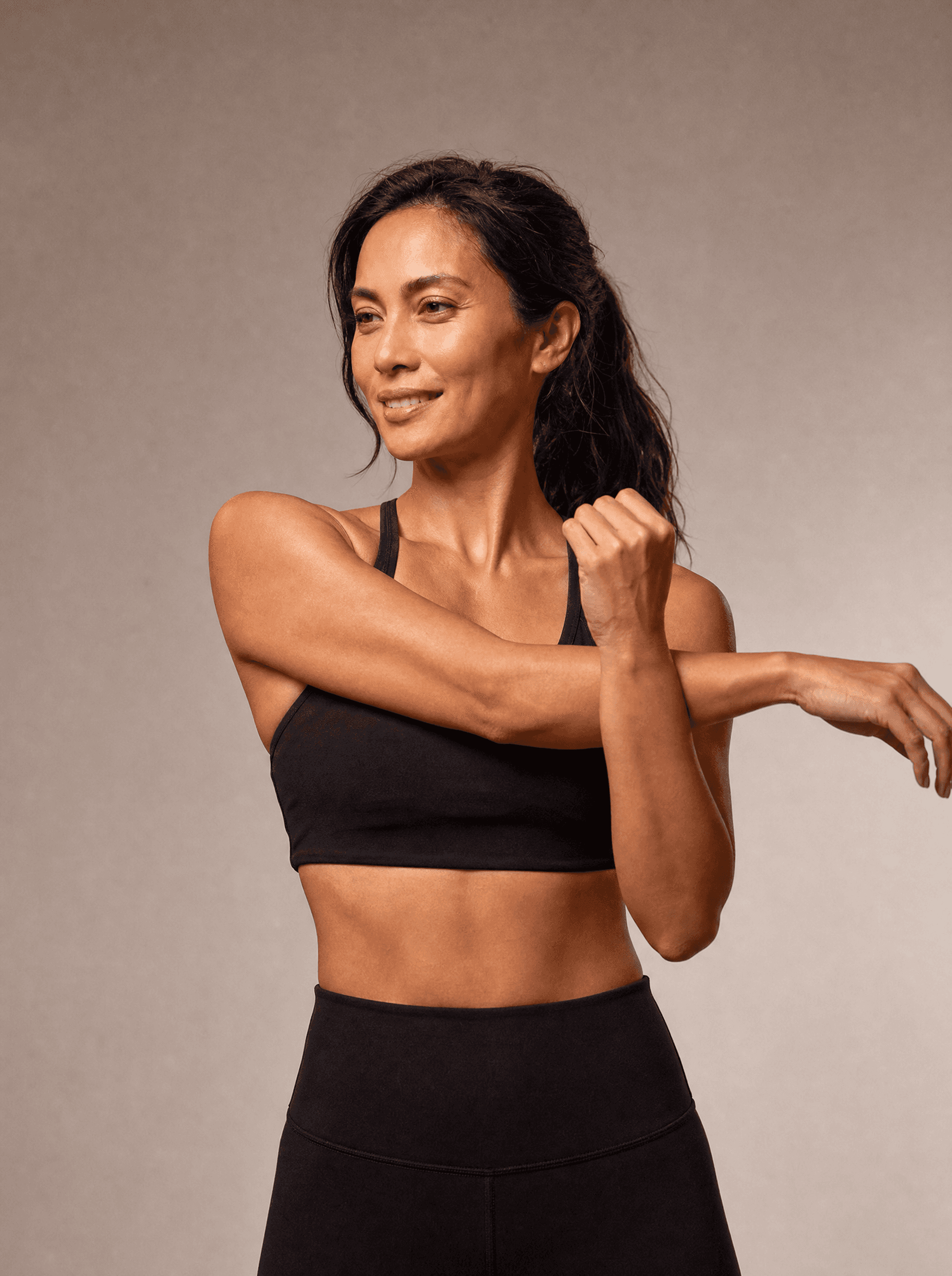 Woman in black athleisure mid-stretch on a warm grey studio backdrop