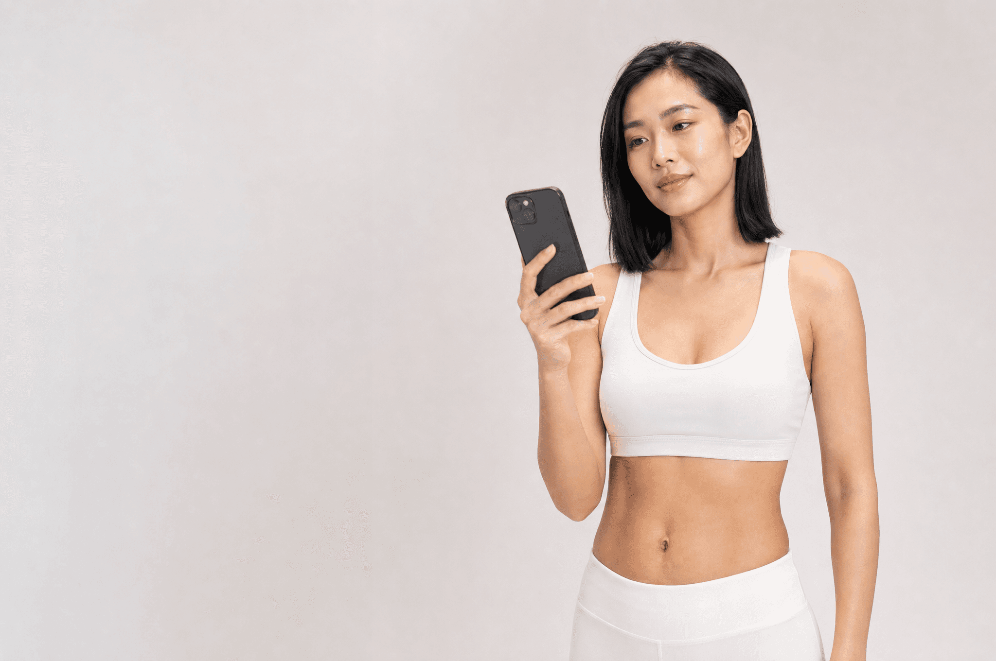 A woman in white athleisure holding a phone up in one hand, glancing at it with a half smile