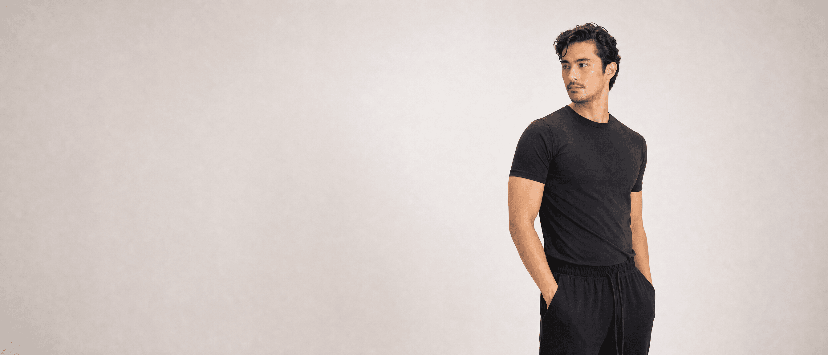 A man in black athleisure standing in a white studio, hands in his pockets, looking off to one side