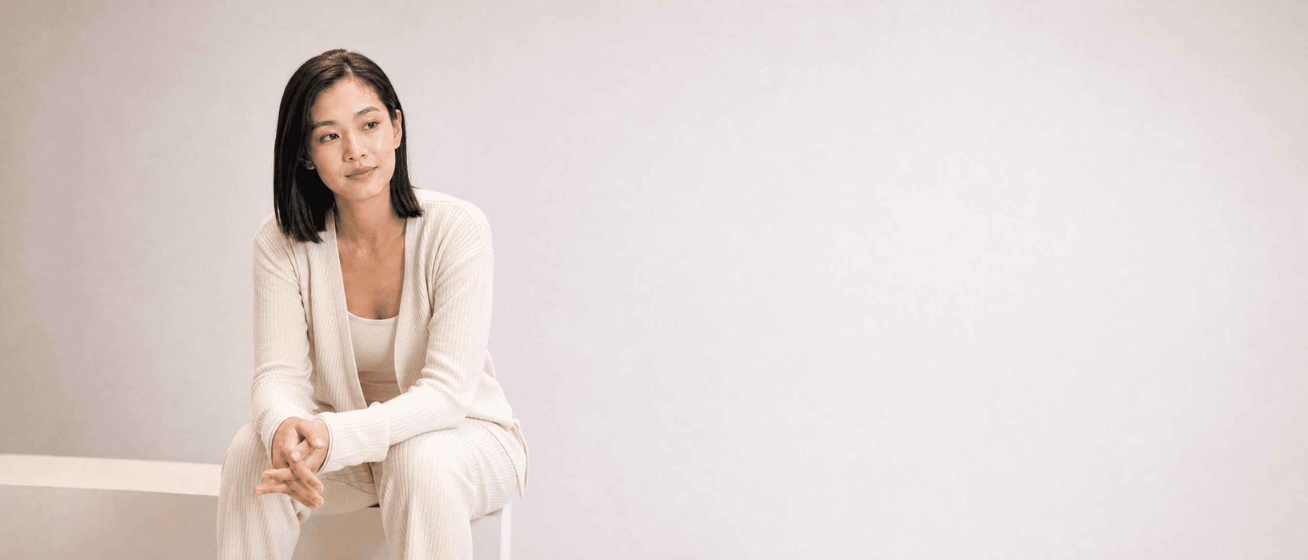 A woman in bone knit loungewear seated on a white plinth, hands clasped on her knee, listening to someone off camera