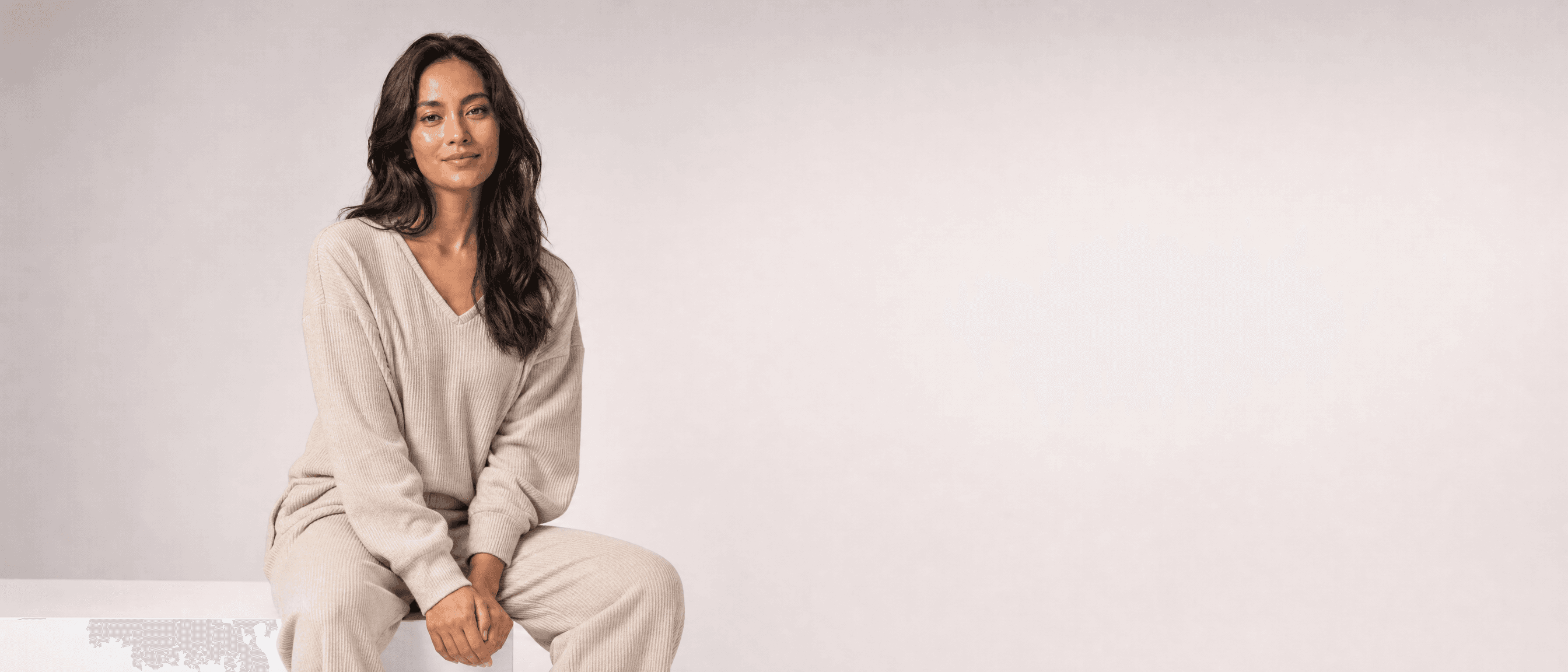 A woman in oat loungewear seated on a white plinth in a white studio, hands folded in her lap, looking at camera