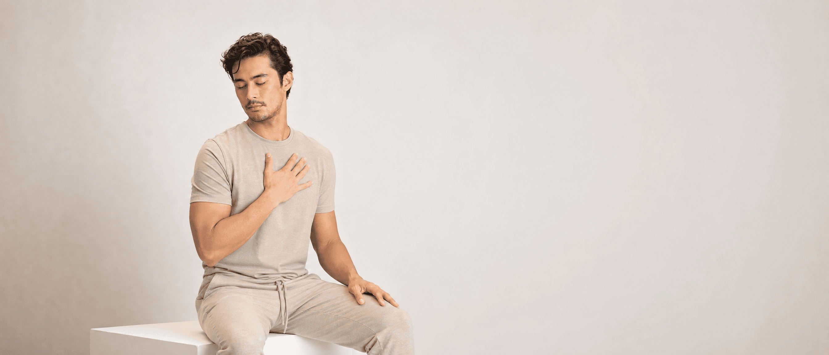 A man in oat loungewear seated on a white plinth, eyes closed, one palm resting on his chest