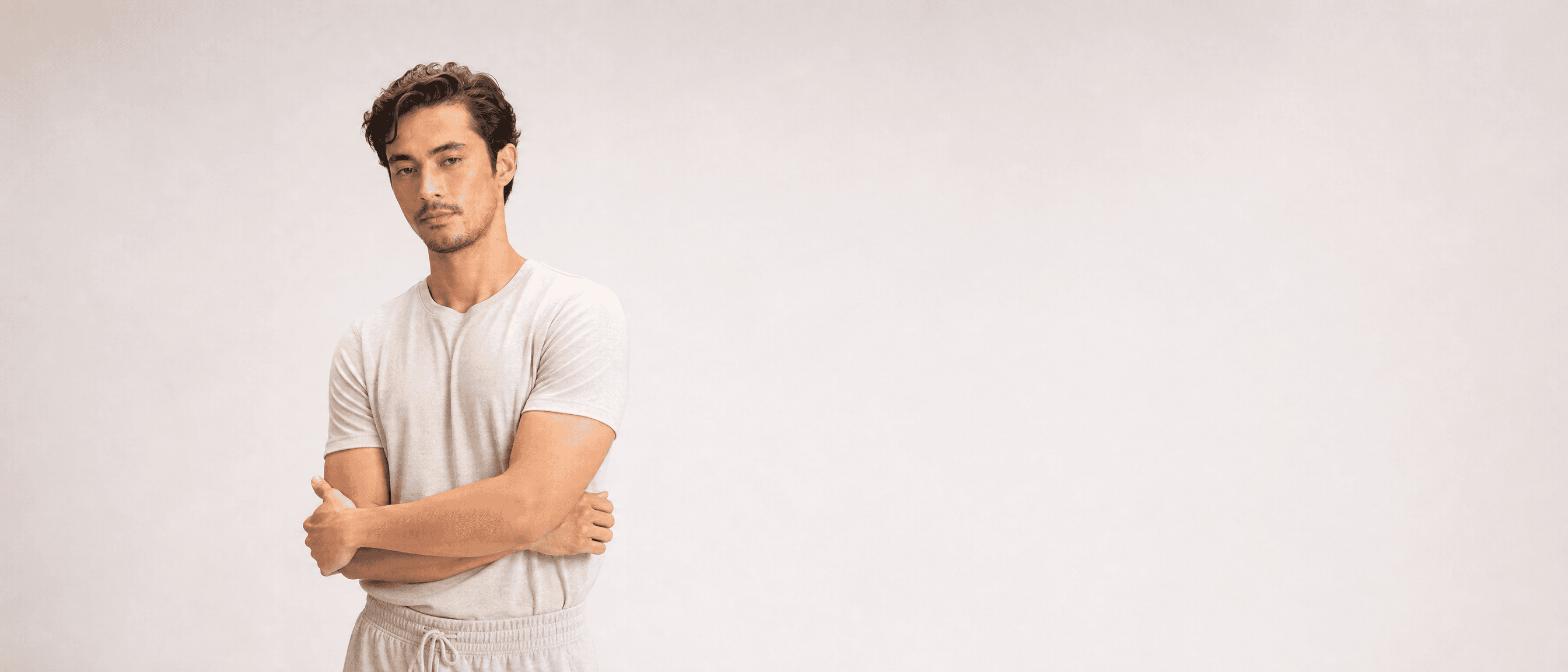 A man in oat loungewear standing in a white studio, arms folded loosely, looking at camera