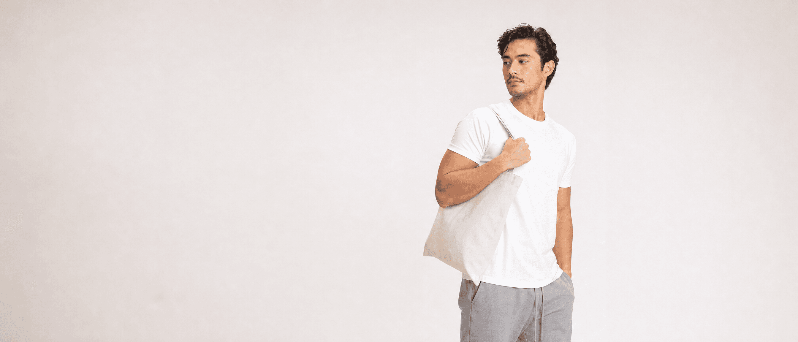 A man in a white tee and grey loungewear with a plain canvas tote over one shoulder, looking off-frame