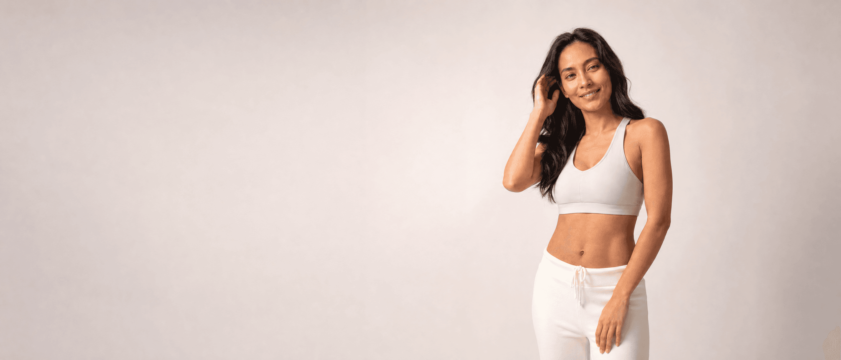 A woman in white athleisure tucking her hair behind her ear, smiling at camera in a white studio