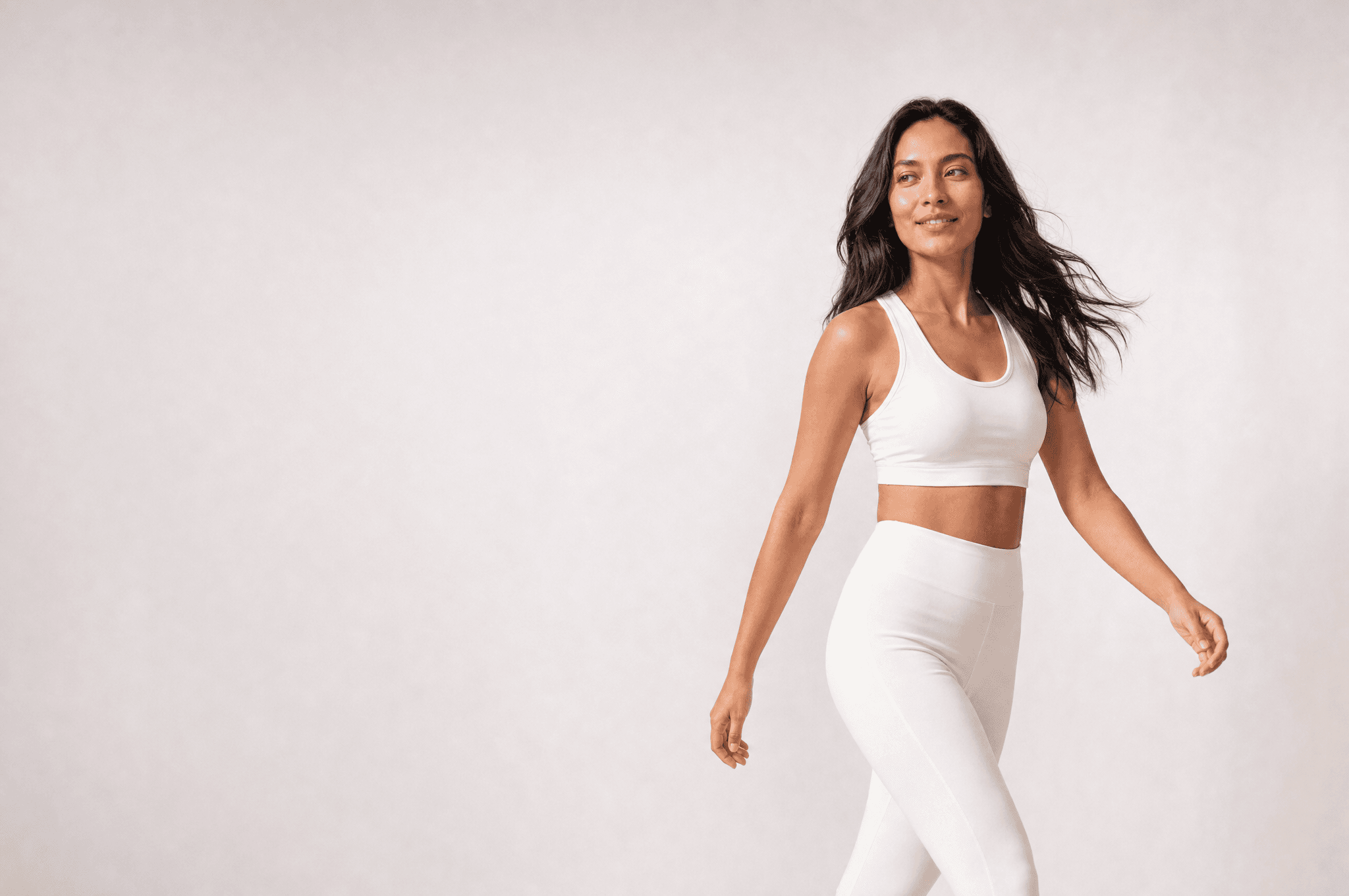 A woman in white athleisure walking across a white studio, hair moving, looking back over her shoulder