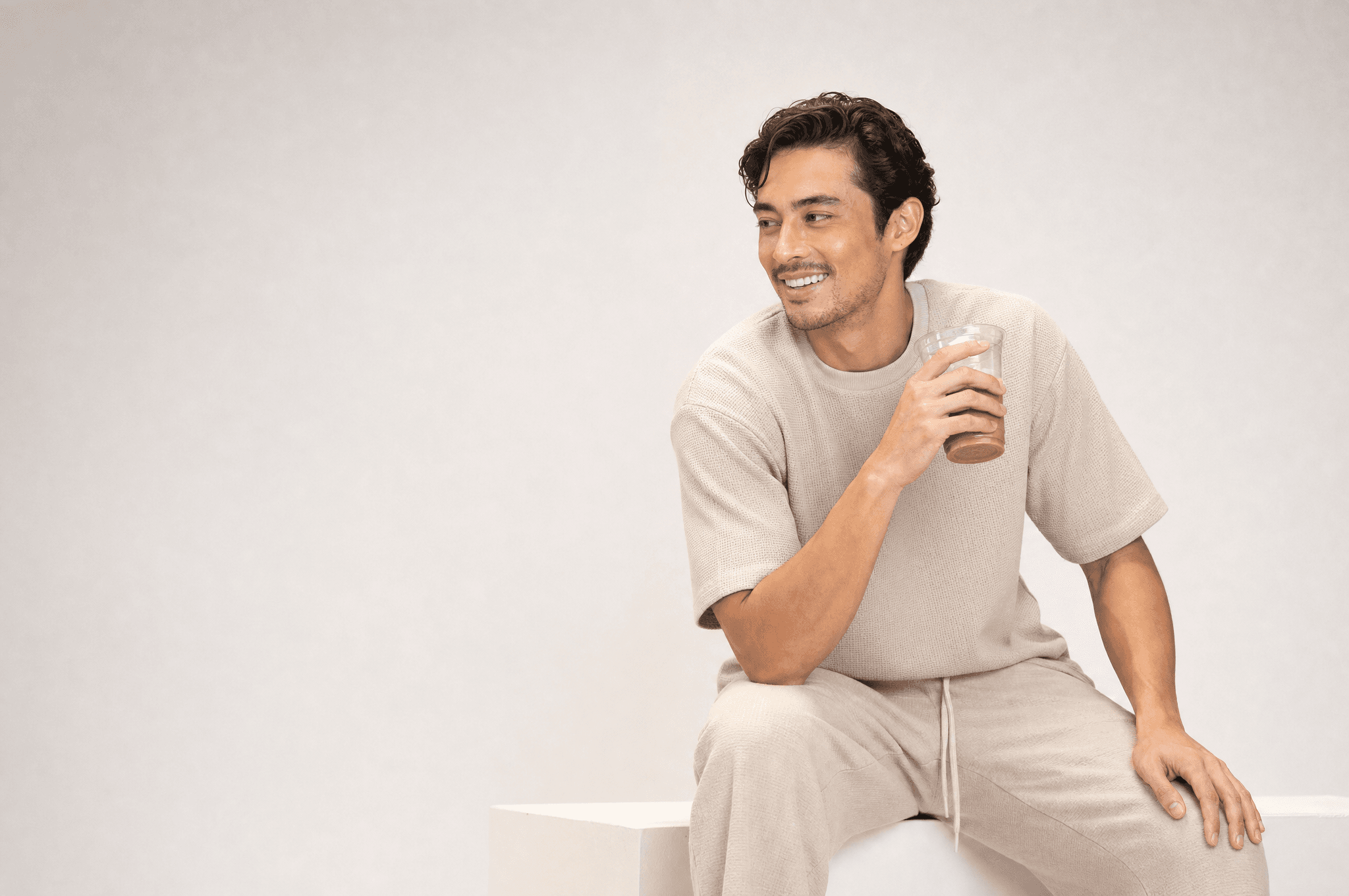 A man in oat loungewear seated on a white plinth, laughing, holding an iced coffee in a clear cup