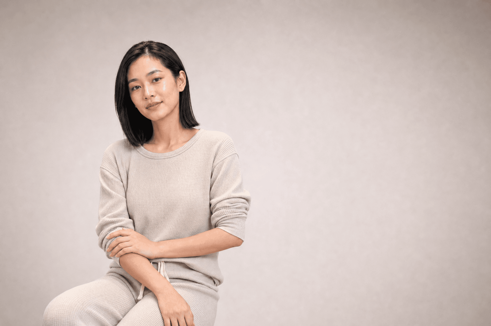 A woman in oat loungewear seated in a white studio, one hand resting on her forearm