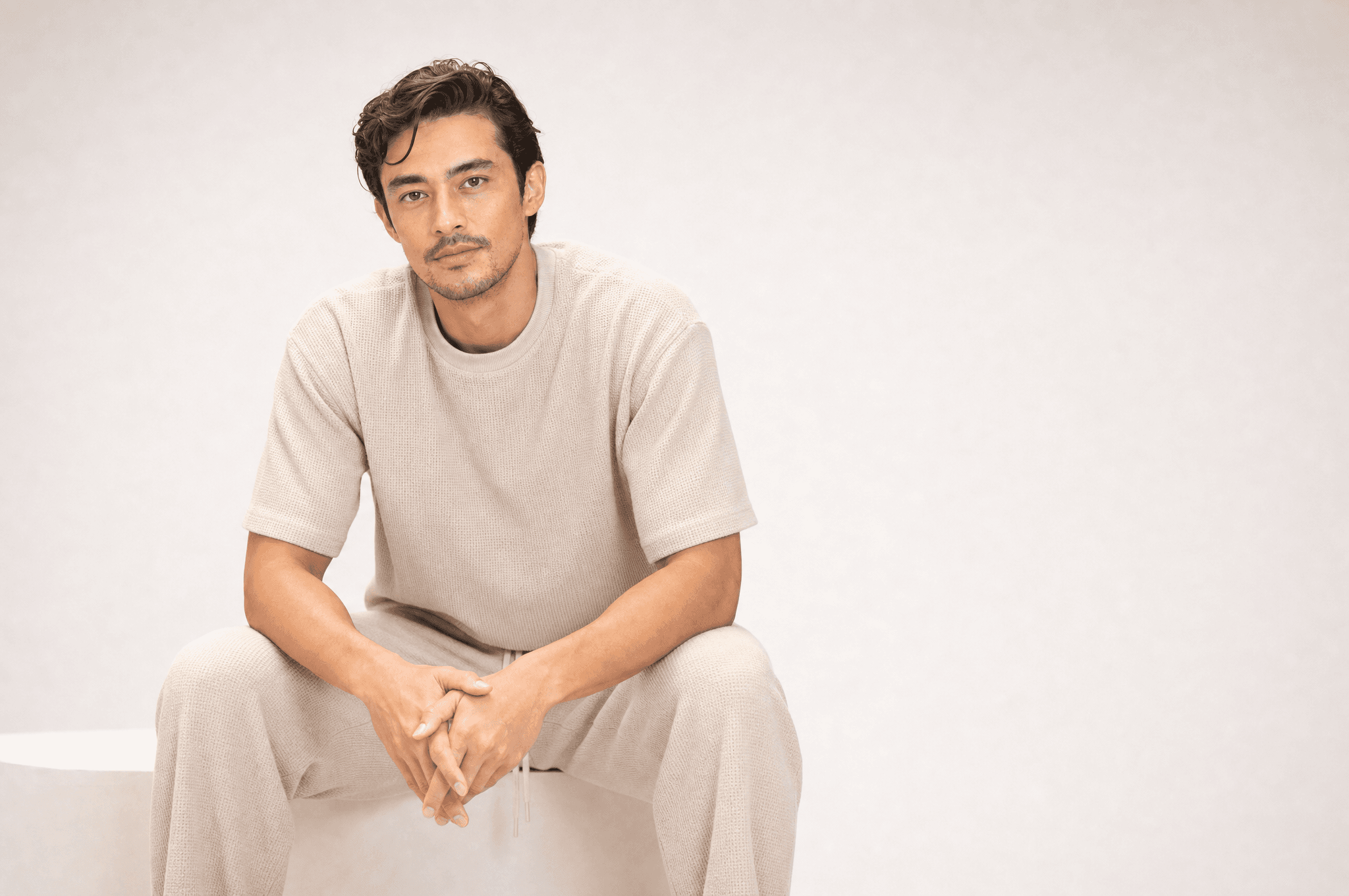 A man in oat loungewear seated on a white plinth, elbows on his knees, hands clasped