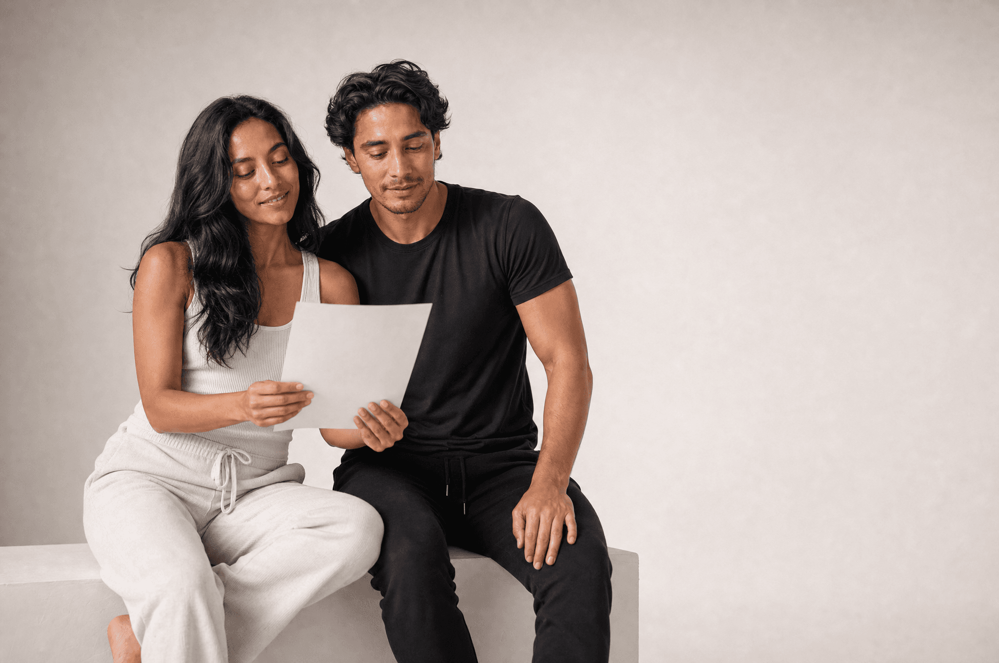 A woman and a man seated on a white plinth reading a single blank sheet of paper together