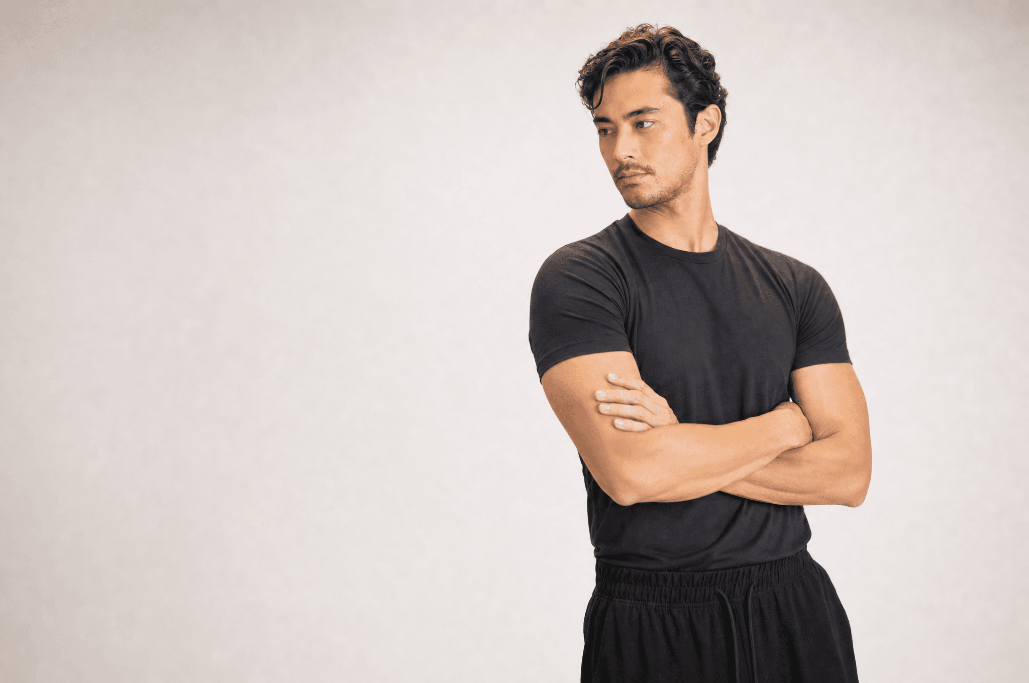A man in a black tee standing in a white studio with his arms crossed, looking to the side