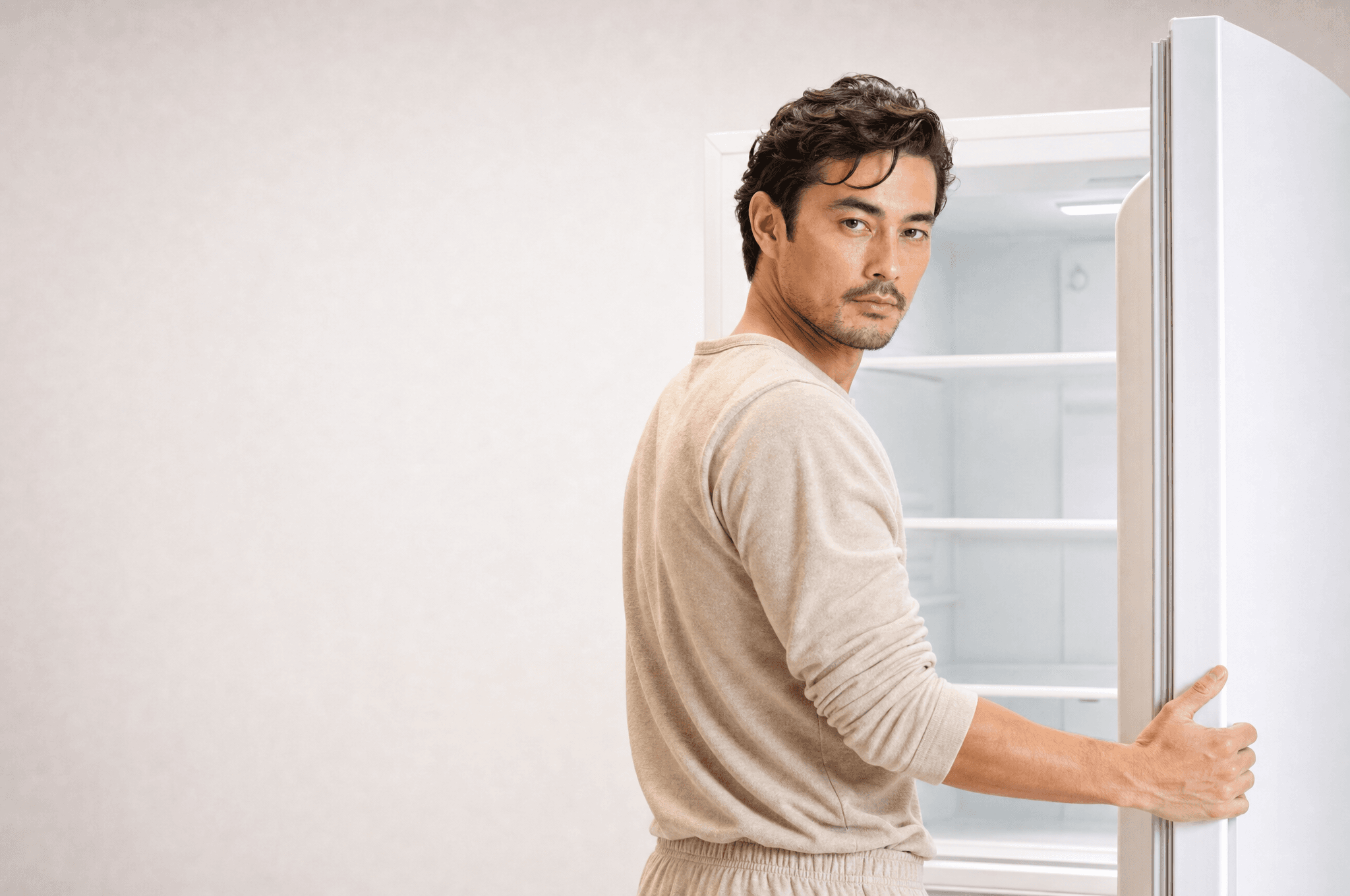 A man in oat loungewear holding open the door of an empty white fridge, looking back at camera
