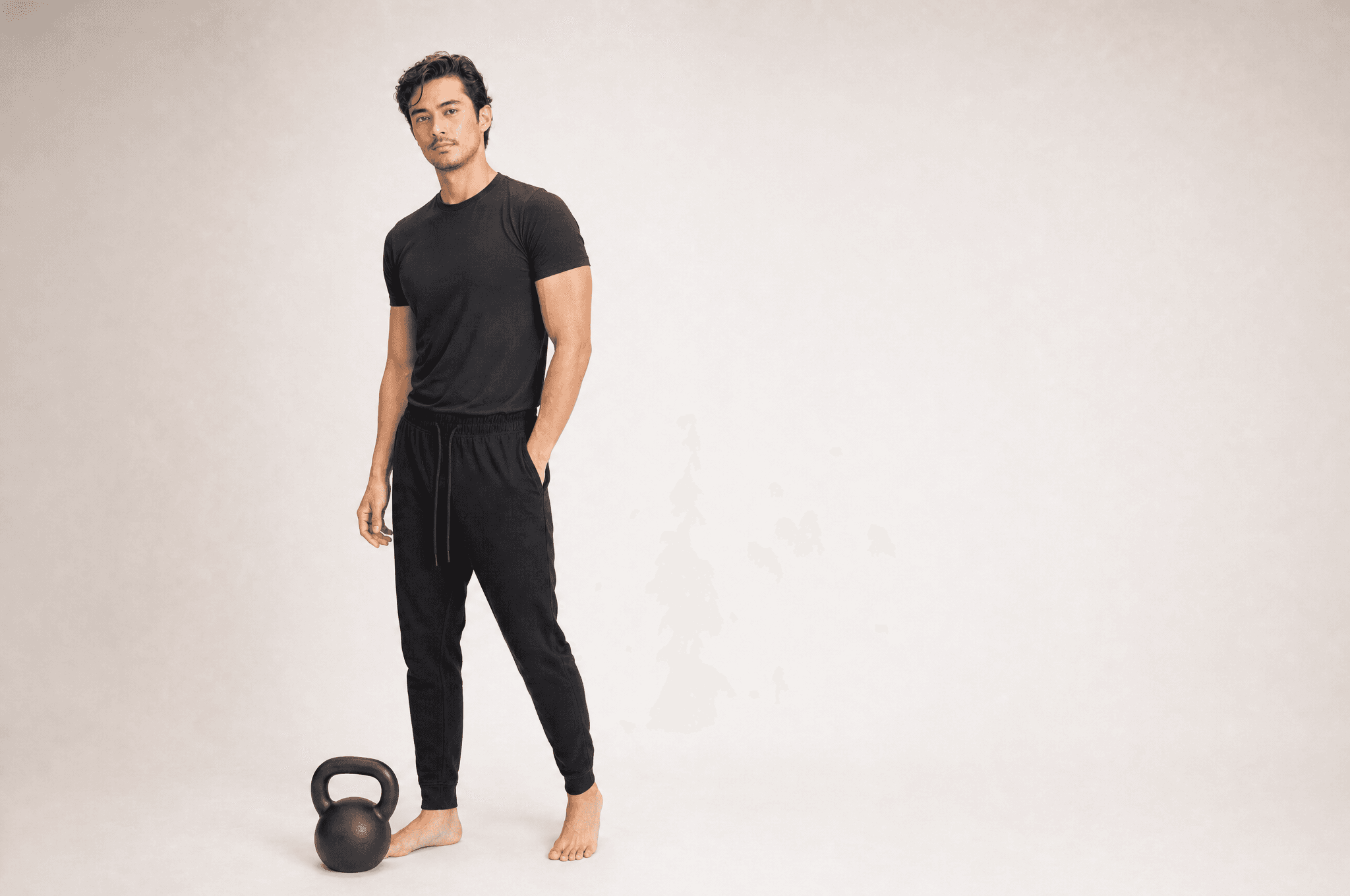 A man in black athleisure standing in a white studio with a black kettlebell at his feet