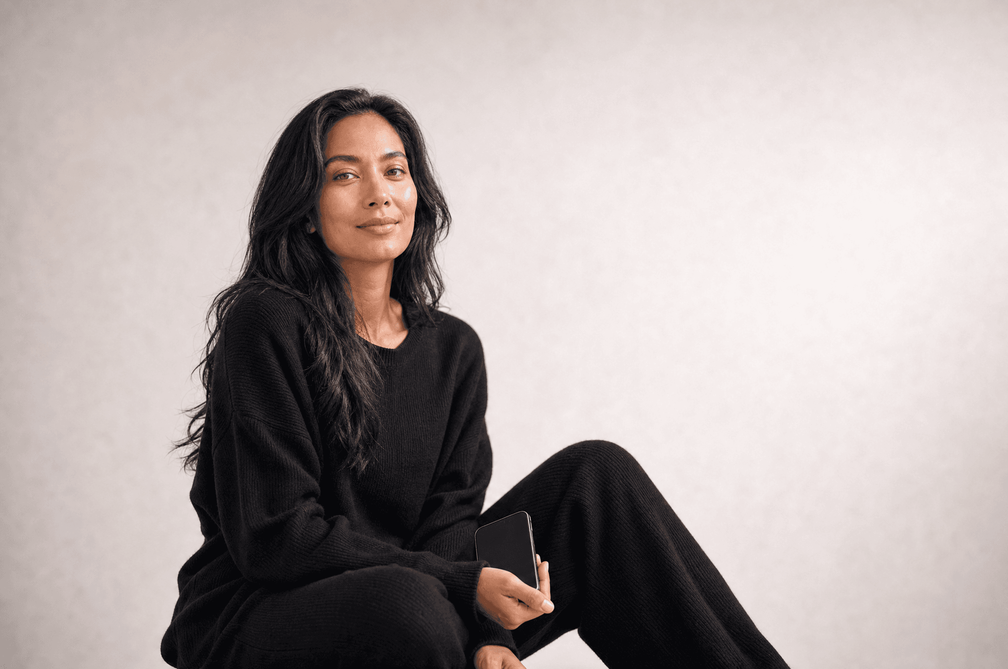 A woman in black loungewear seated in a white studio holding a phone loosely in one hand