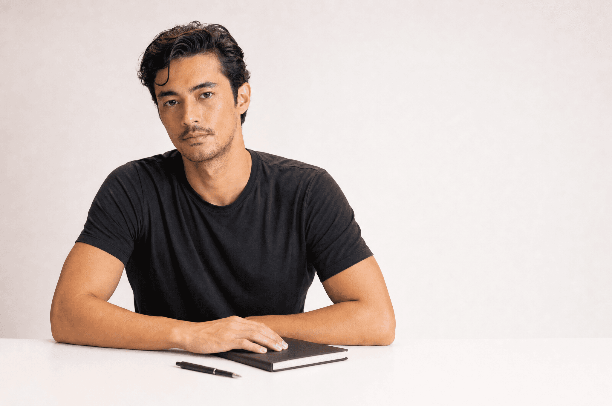 A man in a black tee seated at a white counter with a closed notebook and a pen, looking at camera