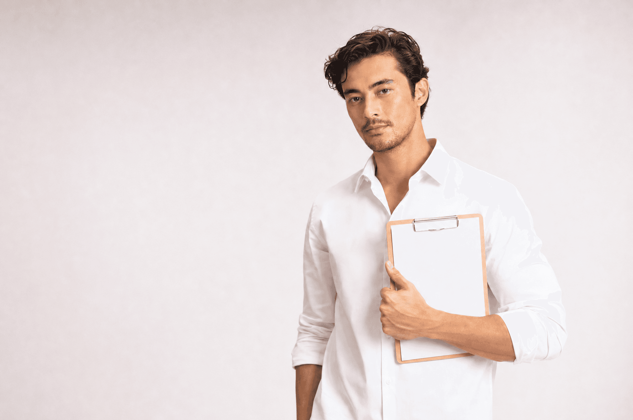 A man in a white collared shirt holding a clipboard with a blank page, looking at camera in a white studio
