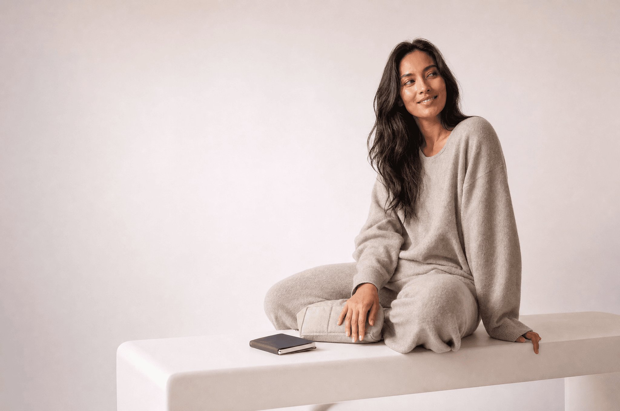 A woman in grey loungewear seated on a long white bench with a small canvas pouch and a notebook beside her