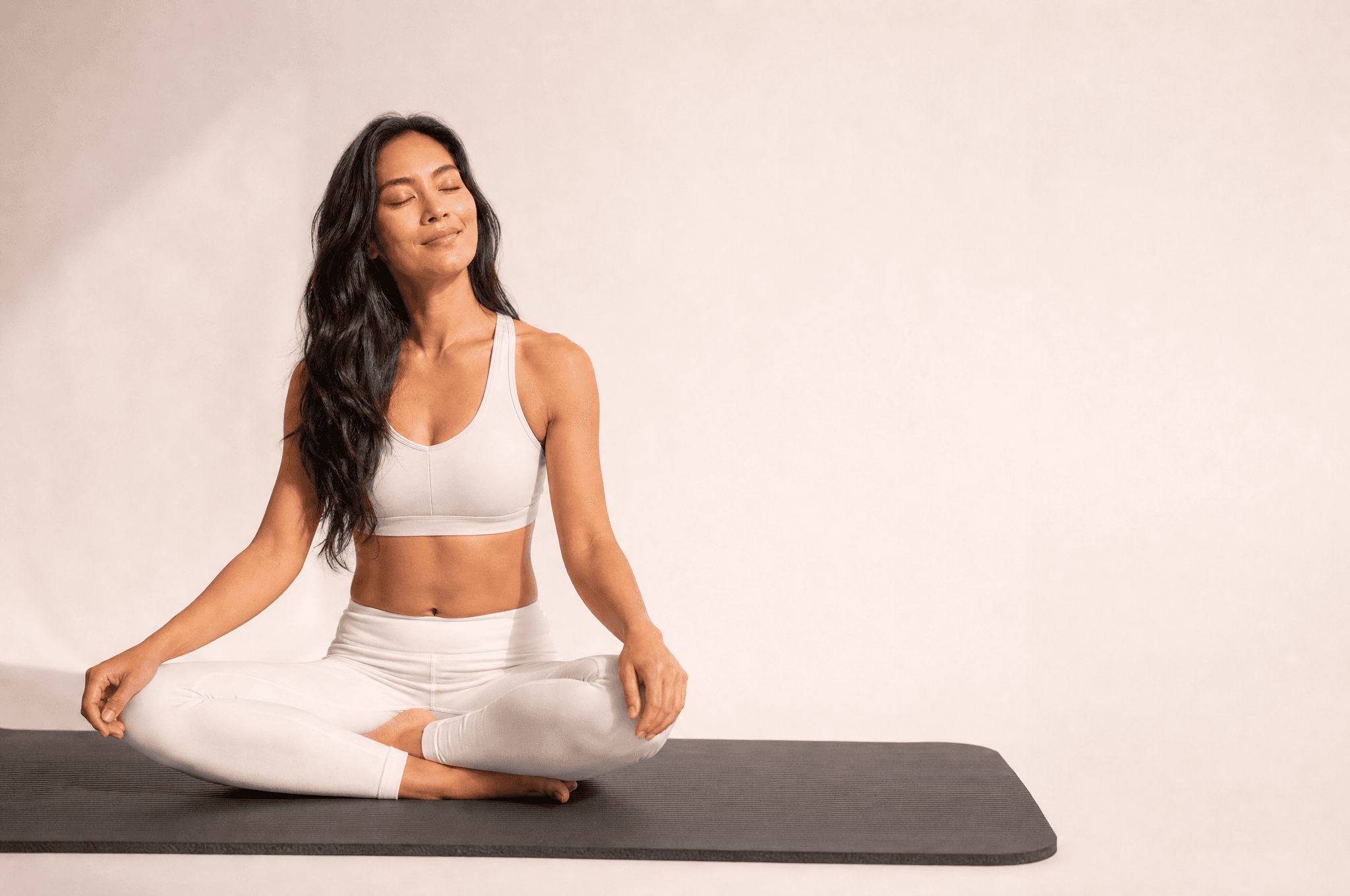A woman in white athleisure seated cross-legged on a grey mat, eyes closed, face turned to the light