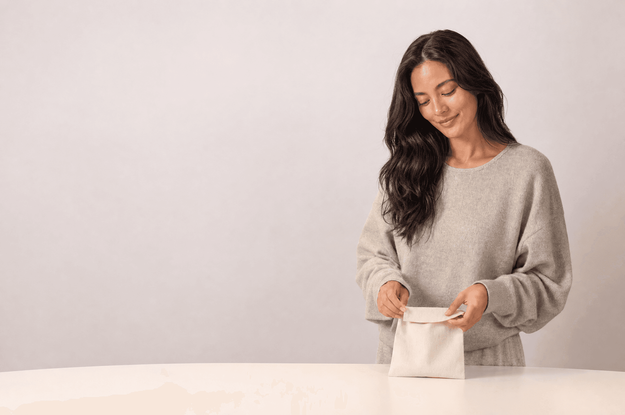 A woman in oat loungewear folding a small canvas pouch closed on a white counter