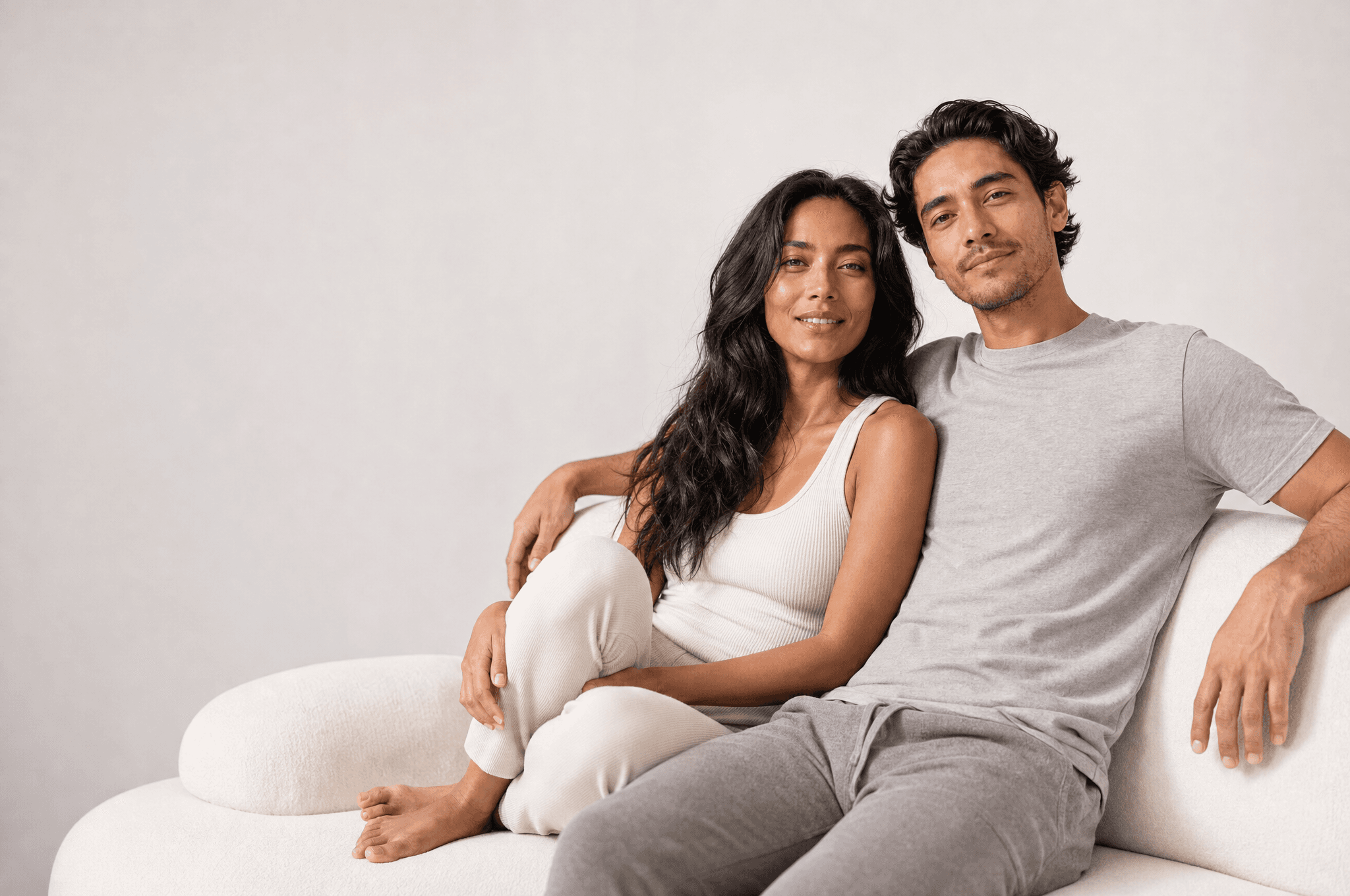 A woman and a man seated close on a low white sofa in a white studio, at ease, looking at camera