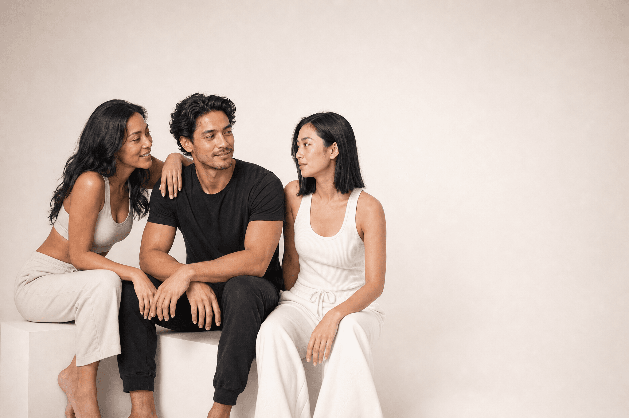 Three people seated together on a low white plinth in a white studio, one leaning in to talk