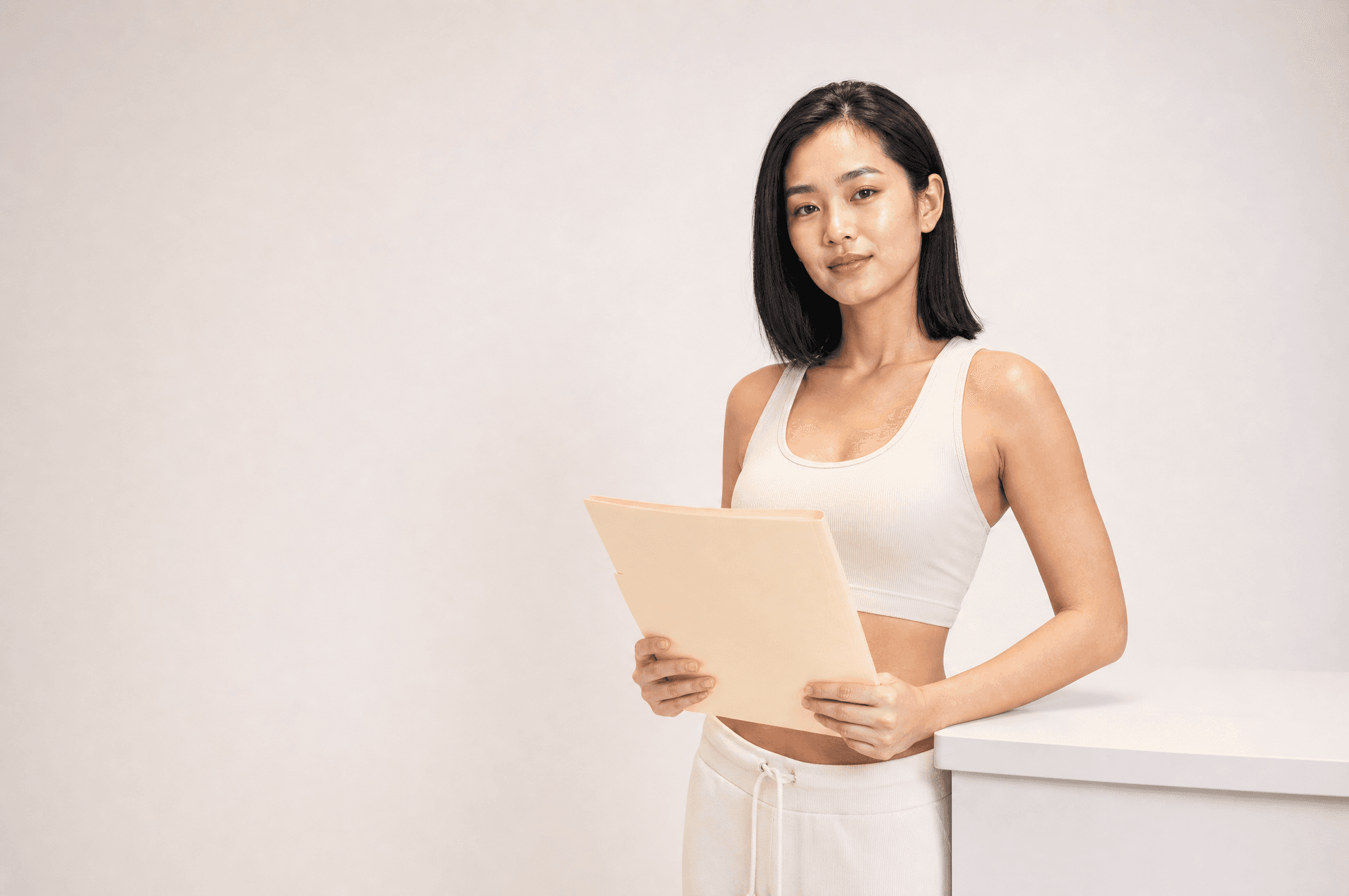 A woman in bone loungewear at a white counter holding a closed folder, looking at camera