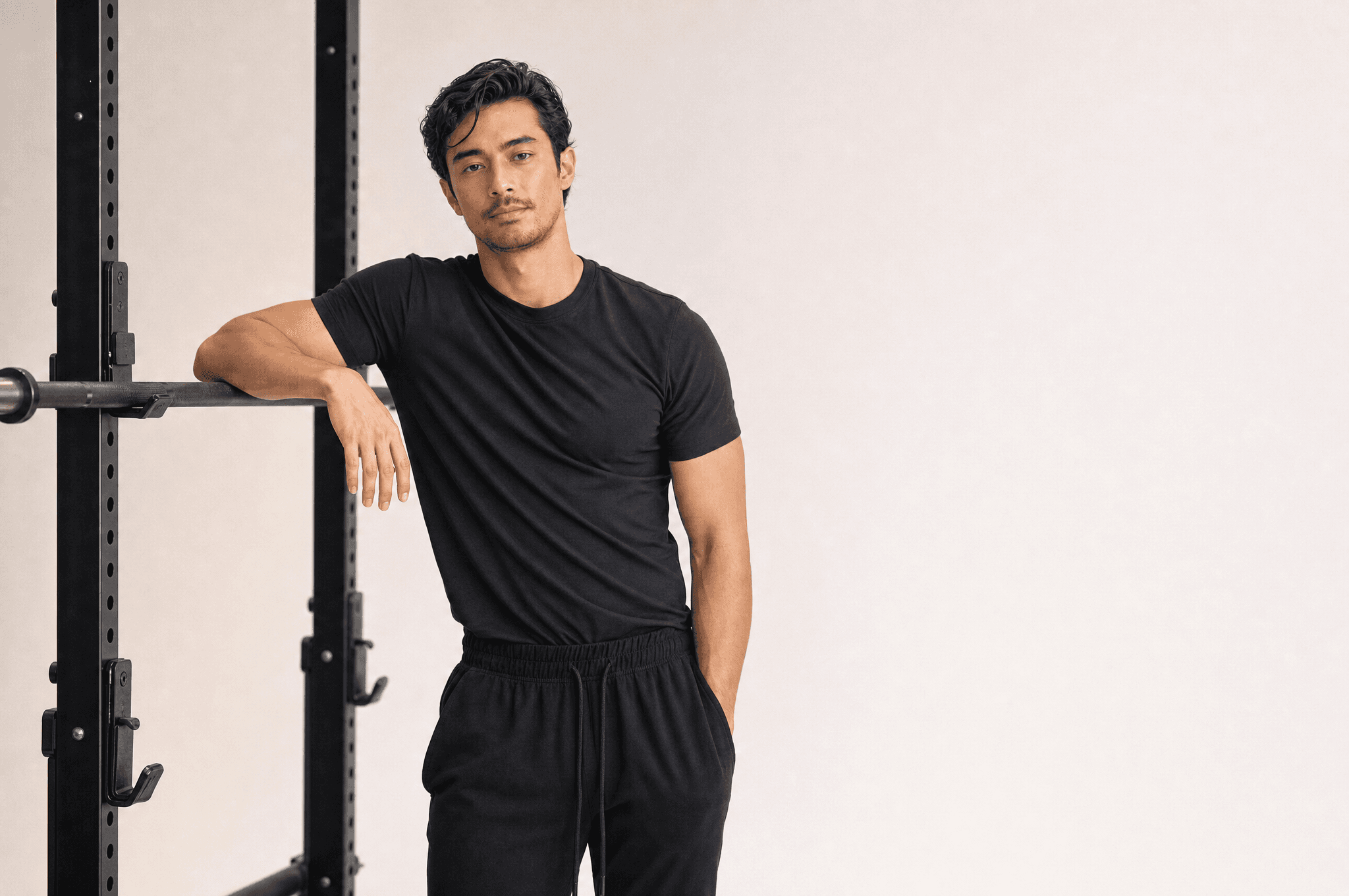 A man in black athleisure resting a forearm on an empty barbell in a squat rack, white studio