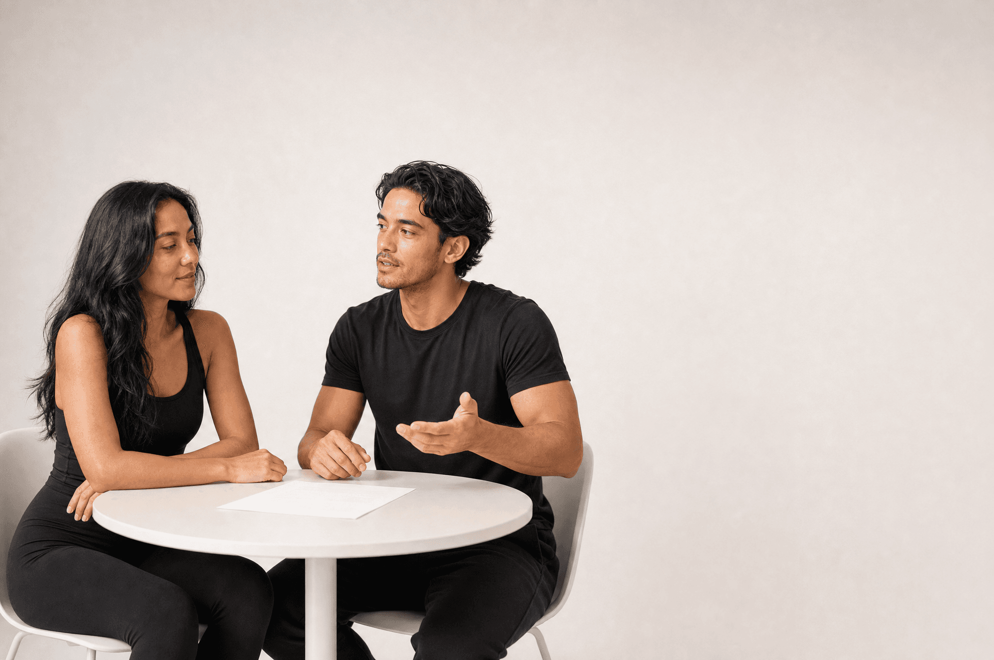 A man and a woman across a small white table, he explaining with an open hand, a blank sheet between them