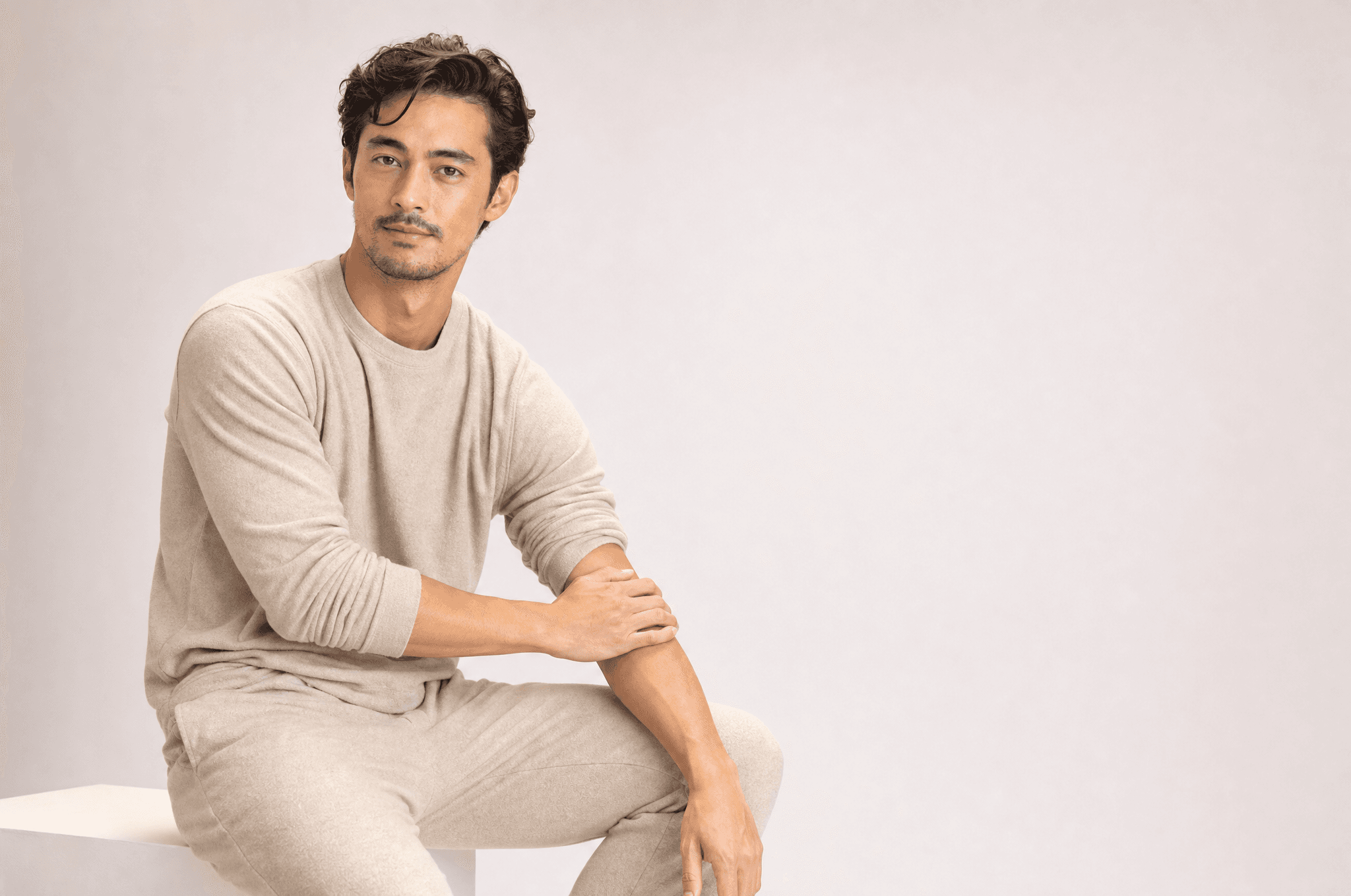 A man in oat loungewear seated on a white plinth, one sleeve pushed up, a hand resting on his forearm