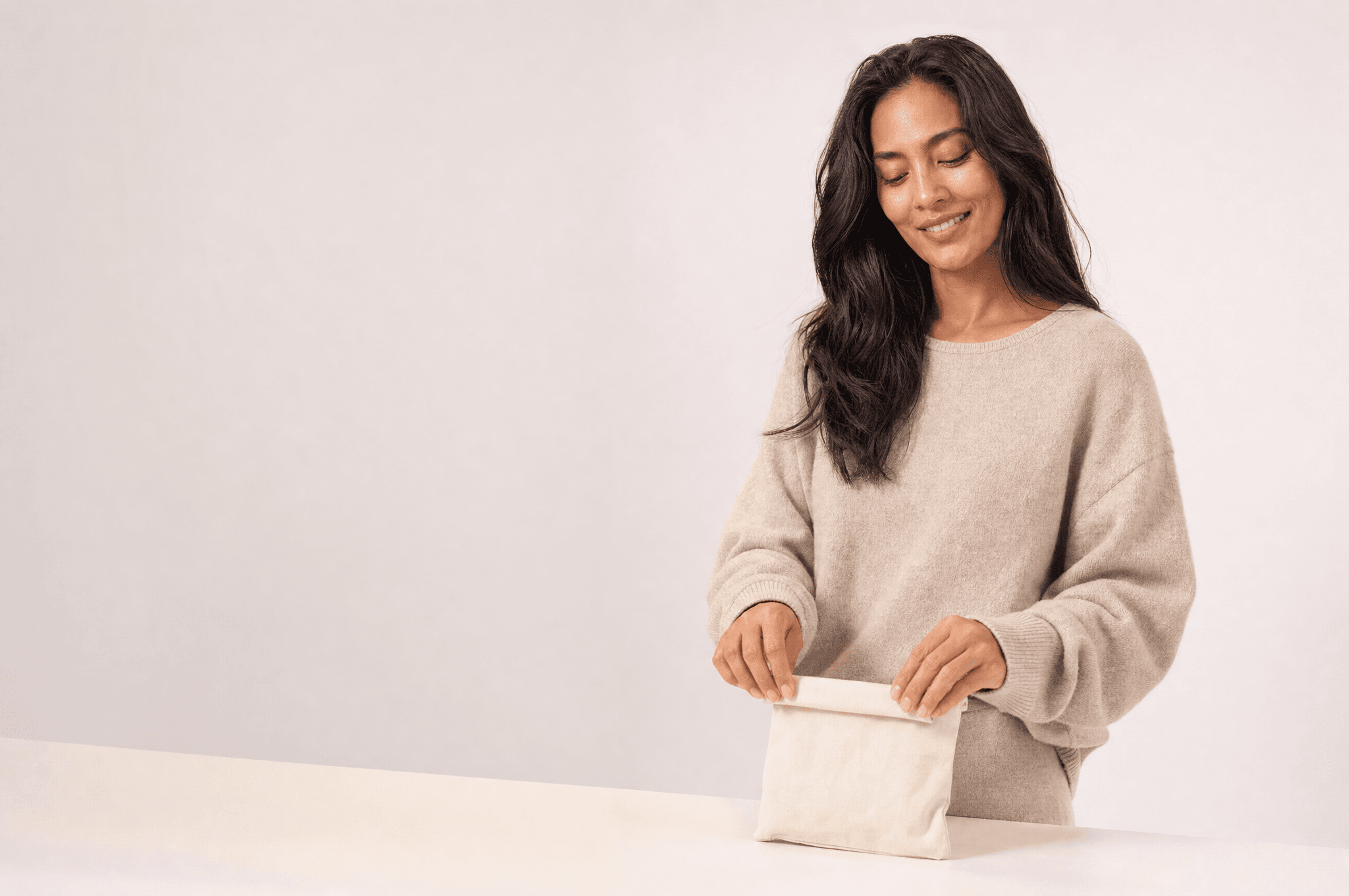 A woman in an oat sweater folding a small canvas pouch closed on a white counter, looking down at her hands
