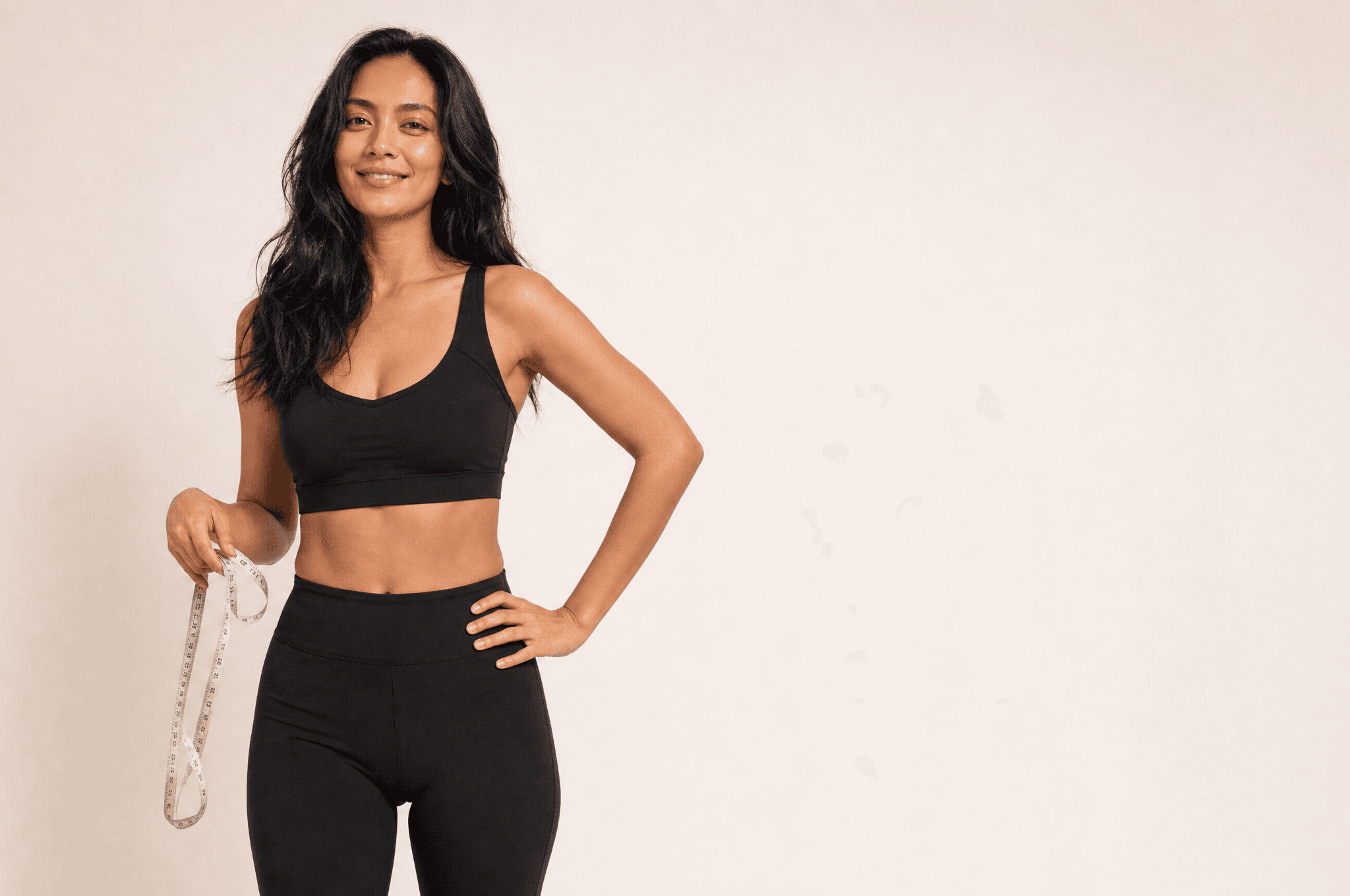 A woman in black athleisure standing in a white studio, a cloth tape measure held loosely in one hand