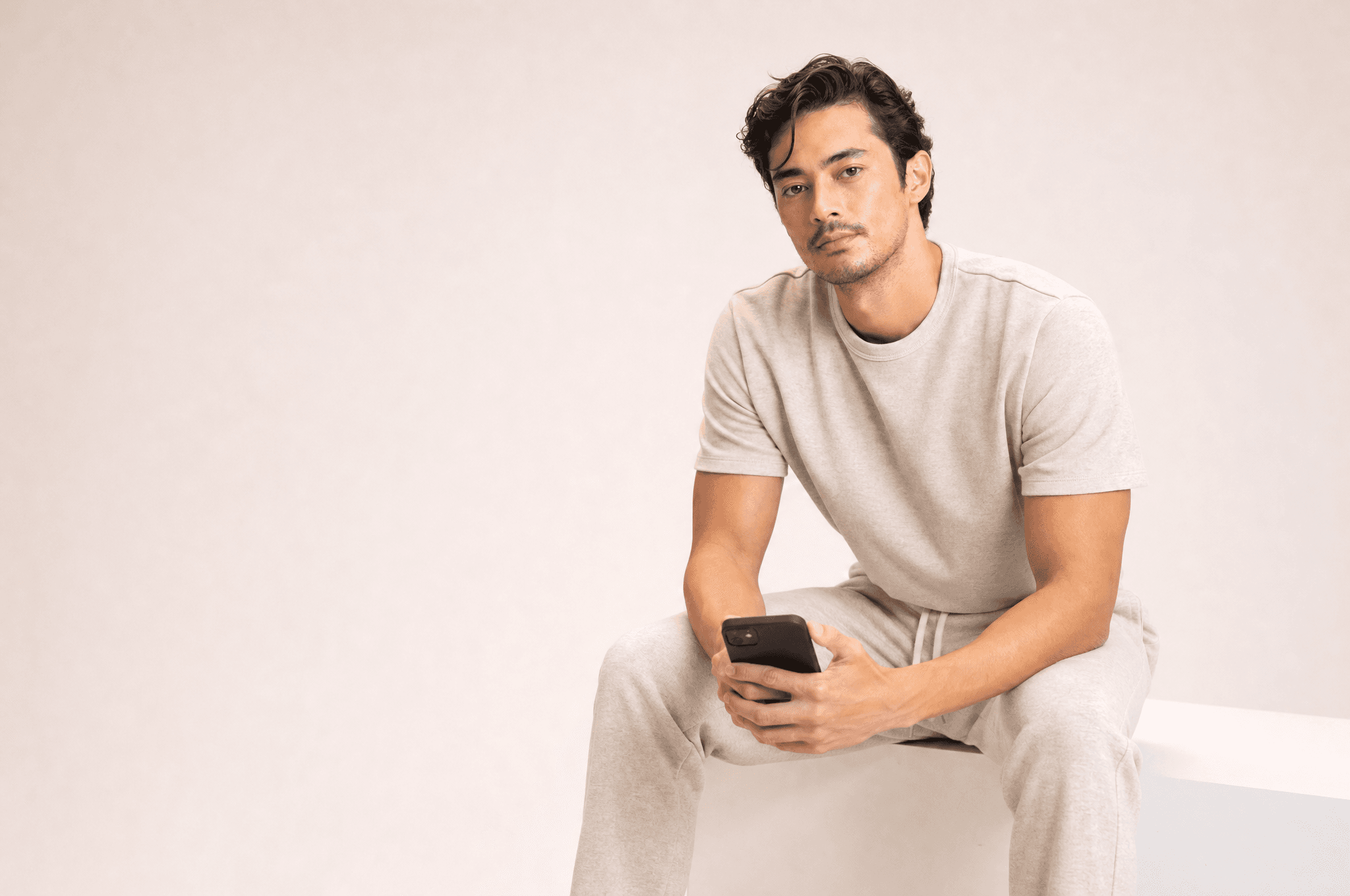 A man in oat loungewear seated on a white plinth holding a phone low in both hands, looking at camera