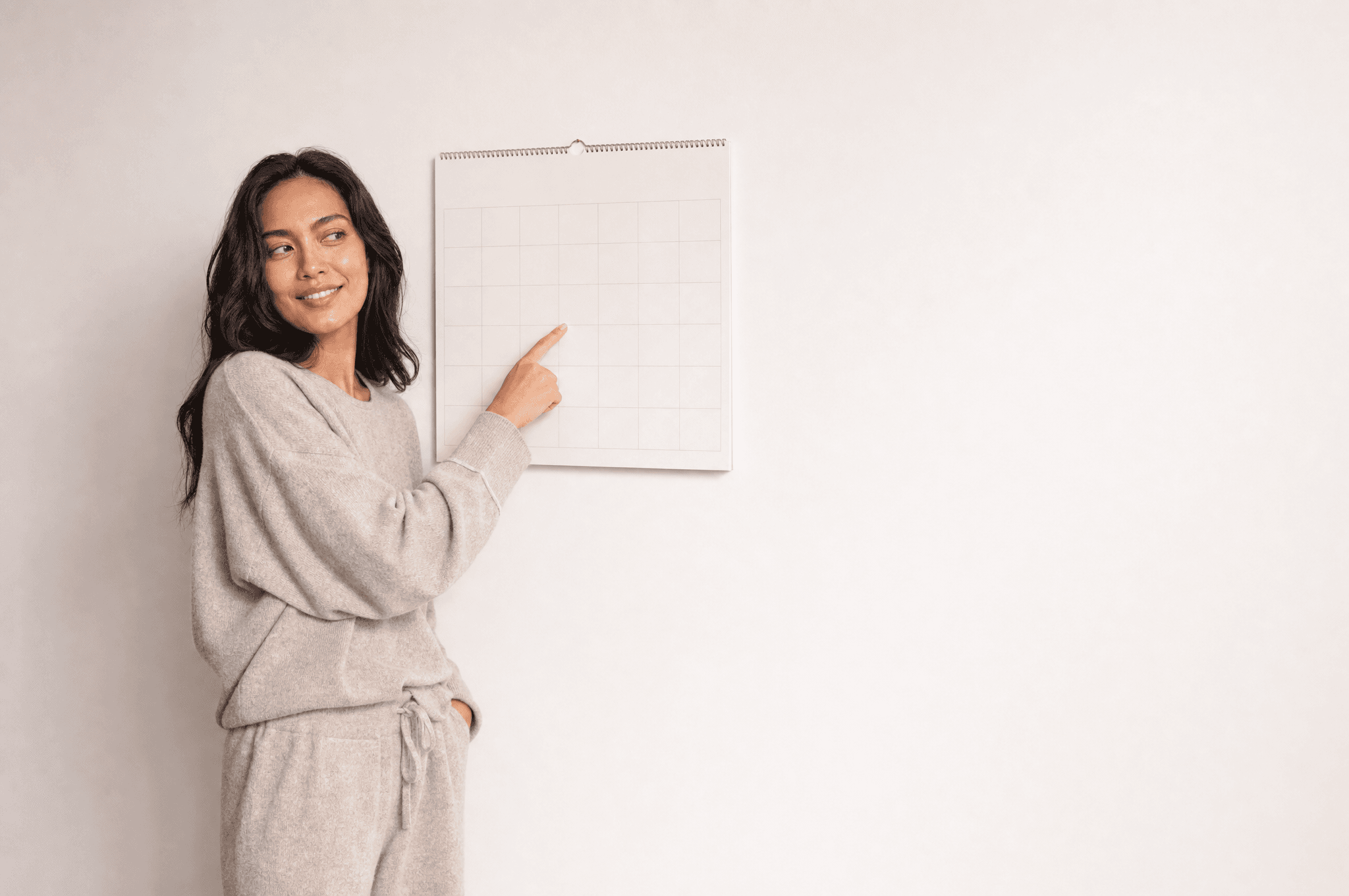 A woman in grey loungewear standing beside a plain wall calendar in a white studio, one finger on a square