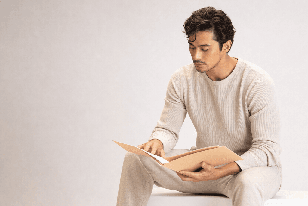 A man in oat loungewear on a white plinth reading an open folder of blank pages across his lap