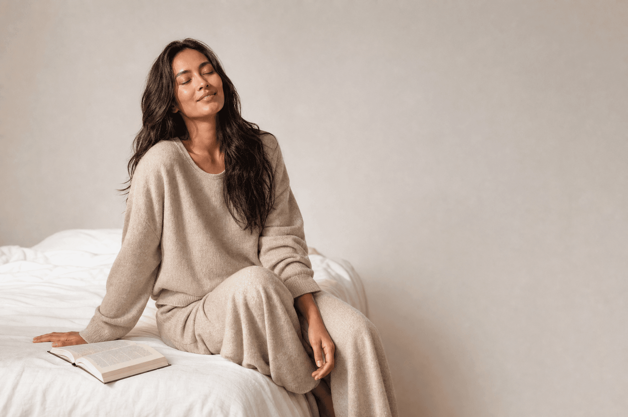 A woman in oat loungewear on the edge of a white bed, eyes closed, an open book face down beside her