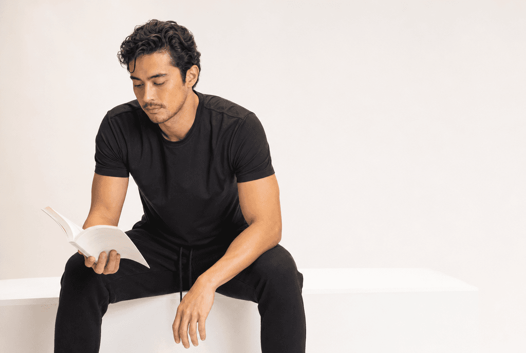 A man in black athleisure on the edge of a white plinth looking down at an open book in one hand