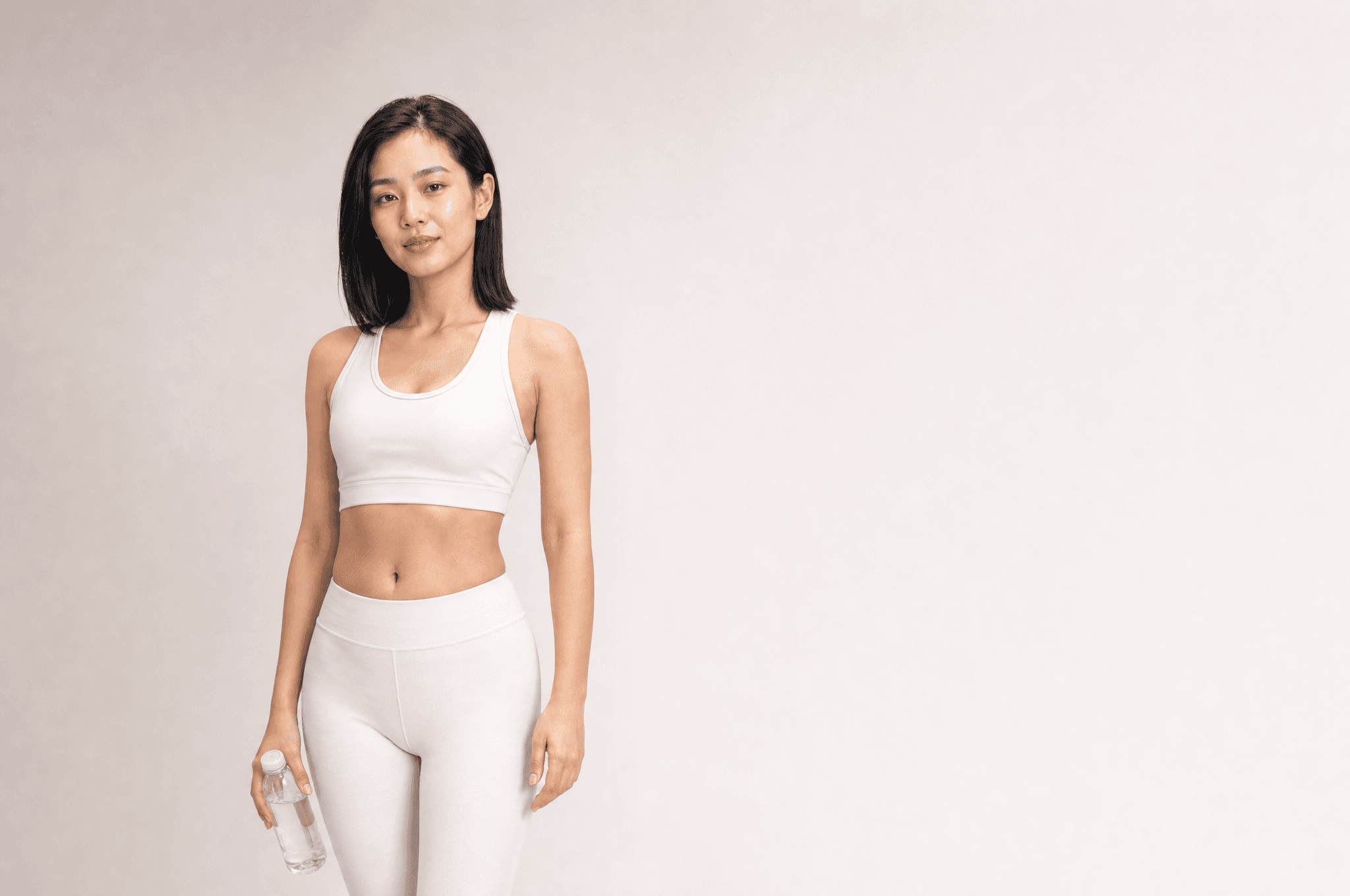 A woman in white athleisure holding a plain clear water bottle low in one hand, looking at camera