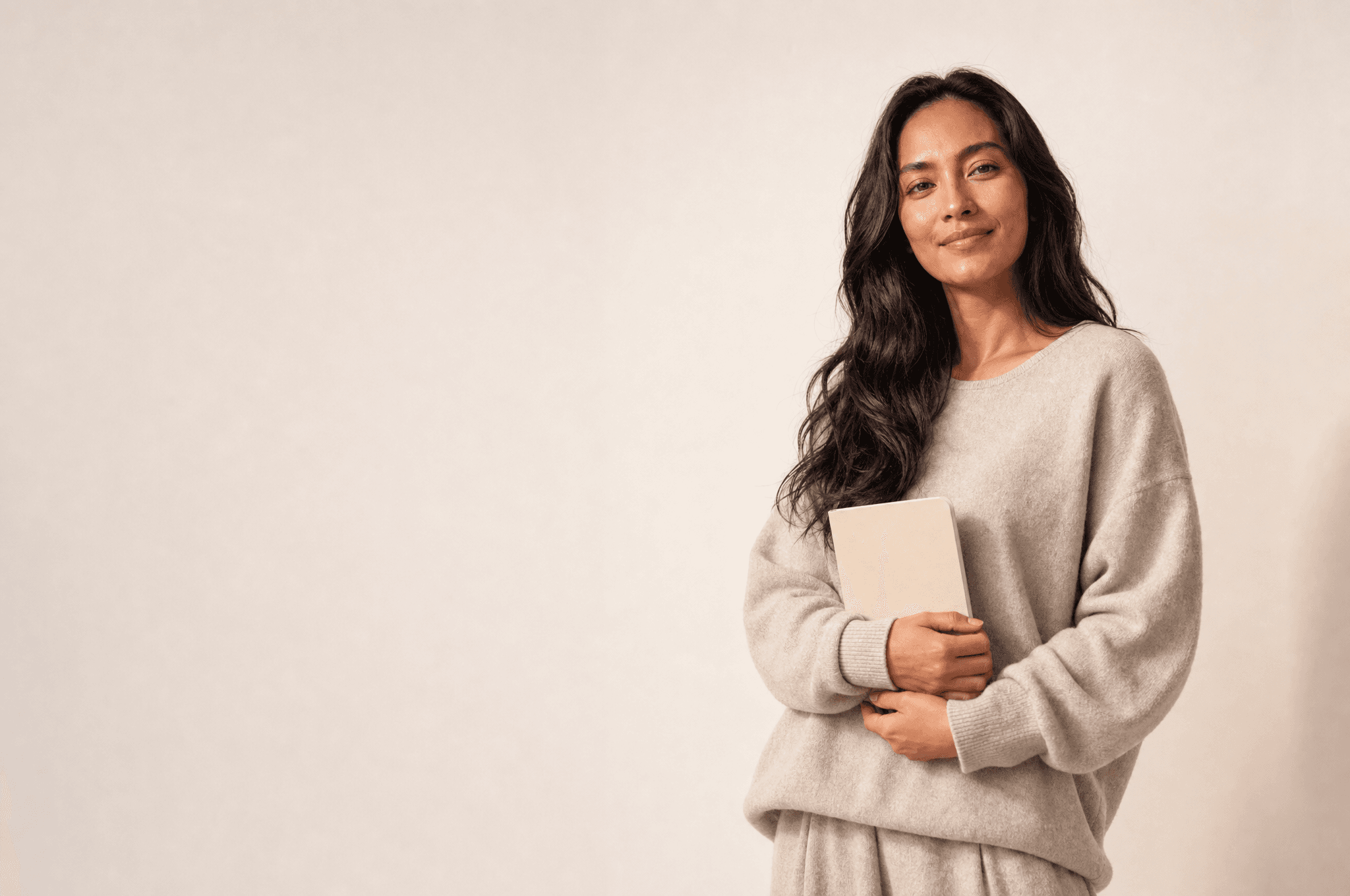 A woman in bone loungewear holding a closed plain notebook against her front in both hands