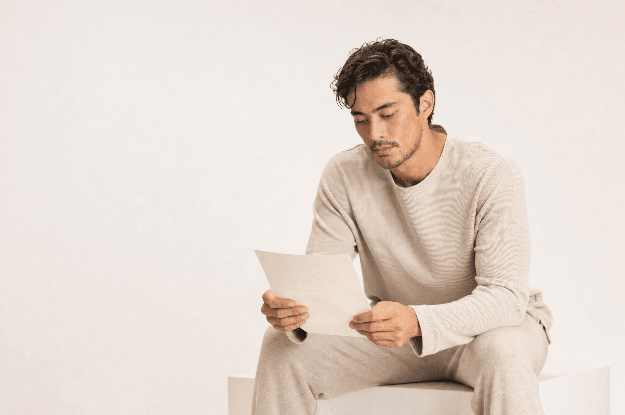A man in oat loungewear seated on a white plinth reading a single folded sheet of paper