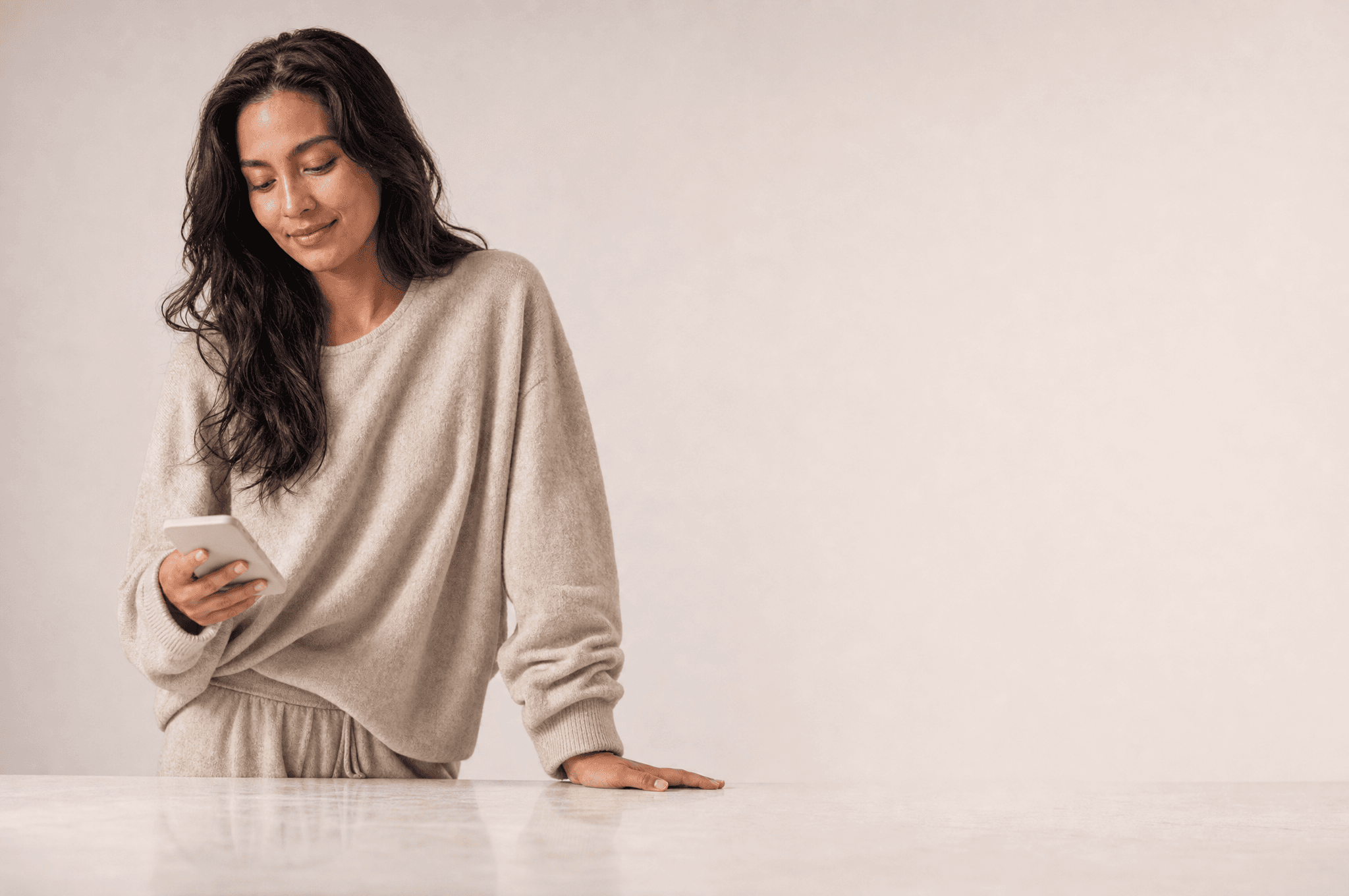 A woman in bone loungewear at a white kitchen counter looking down at a phone held low in one hand
